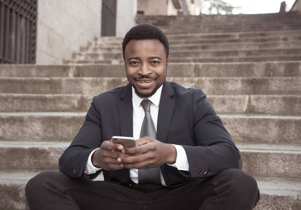快乐的非裔美国商人在手机上查看短信，社交网络坐在城外楼梯上，在人们技术在线连接和企业家创意商业理念上。