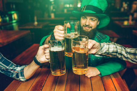 在酒吧里, 穿着绿色圣帕特里克西服的年轻人与朋友坐在桌旁的照片。他们把一杯啤酒放在一起