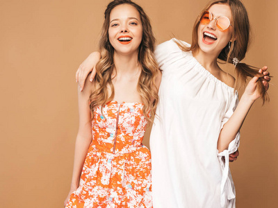 两个年轻美丽微笑的时髦女孩穿着时髦的夏装。 无忧无虑的女人在演播室的黄金背景上摆姿势。 太阳镜正面模特
