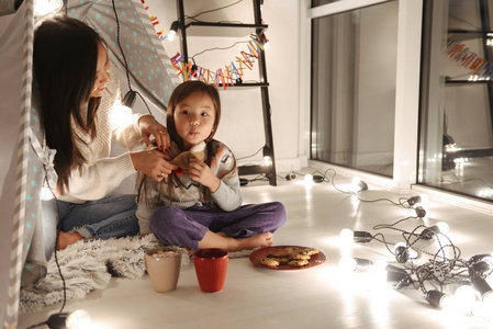 一个年轻的亚洲女人和她的女儿小女孩坐在地板上吃饼干和可可的照片。 圣诞概念。