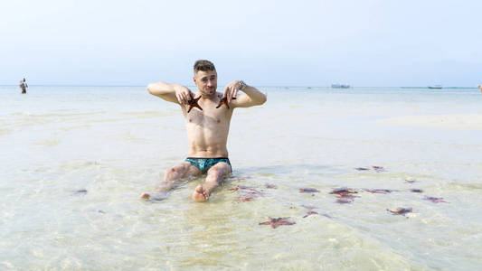旅行男孩正坐在大海上，带着红色的星星鱼在清澈的水中的PhuQuoc岛上拍摄有趣的照片