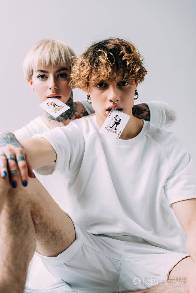 头发卷曲的男人和纹身的女孩嘴里拿着扑克牌,被隔离在灰色上