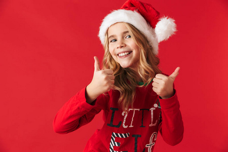 快乐的小女孩戴着圣诞帽，站在红色背景下，竖起大拇指