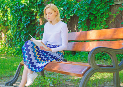 终极最佳图书列表。女金发女郎在公园看书时休息放松。女孩坐板凳放松与书, 绿色自然背景。阅读文学作为爱好。喜欢看书的女孩