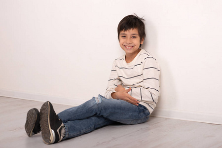 一个穿着白色衬衫和牛仔裤的男孩坐在地板上靠墙休息的摄影棚