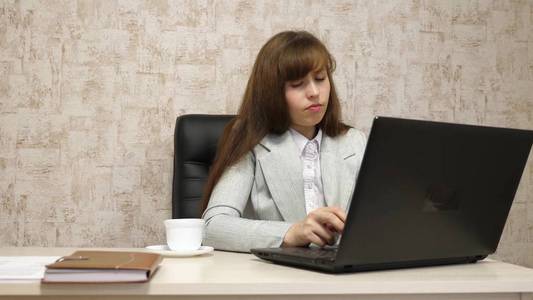 美丽的女孩坐在办公室的椅子上, 在笔记本电脑上打字, 喝白杯咖啡。在电脑工作的年轻女商人。女孩是在笔记本电脑上聊天