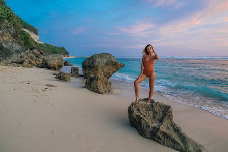 苗条的女孩在泳衣站在大石头在美丽的日落的野生海滩背景