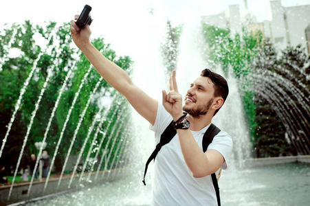 穿着白色 t恤的乐观的积极男子在喷泉附近拿着相机自拍时表现出平和的姿态