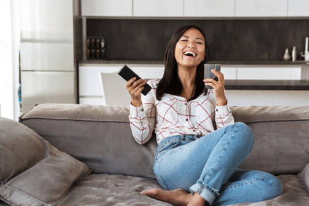 20岁的快乐亚洲女人在舒适的公寓里坐在沙发上拿着遥控器和杯子的照片