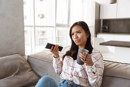 20岁的亚洲美女在舒适的公寓里坐在沙发上拿着遥控器看电视的照片