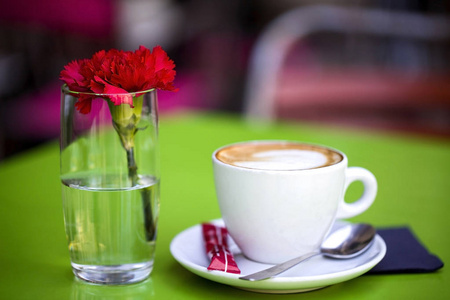 一个白色的杯子在茶托上，热咖啡在绿色的桌子上，在街边的咖啡馆旁边，是一个带有新鲜康乃馨的杯子。 宏观照片模糊背景。