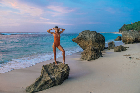 穿着泳衣的瘦身晒黑的女孩, 在日落的海滩背景下, 举起双臂站在大石头上