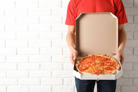 年轻人用打开的披萨盒靠近白色砖墙，空间为文字。提供食品服务。