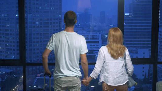 晚上, 在全景窗户的摩天大楼背景下, 有魅力的夫妇女人和男人拿着一个手提箱