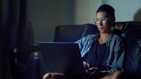 年轻英俊的亚洲男人眼镜与反射使用他的笔记本电脑, 坐在晚上的房间里。不知情