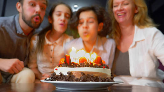 快乐的微笑的男孩吹蜡烛在她的生日蛋糕上。孩子们被他们的家人包围。带蜡烛的生日蛋糕