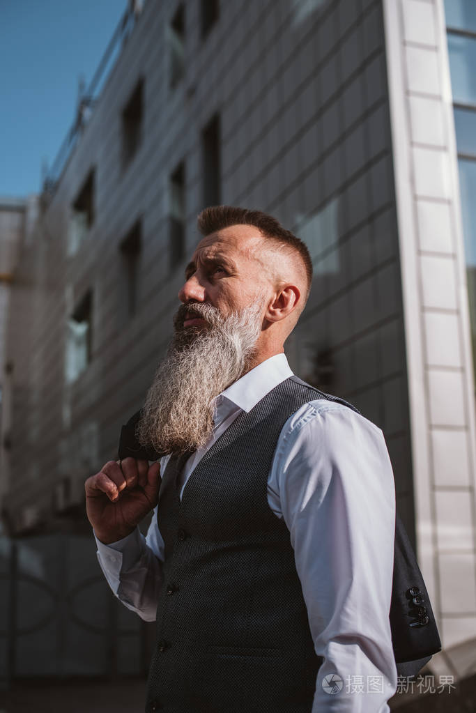 在阳光明媚的白天在现代建筑背景下身着户外西装的英俊胡须成熟男人的