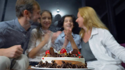 快乐的微笑的男孩吹蜡烛在她的生日蛋糕上。孩子们被他们的家人包围。带蜡烛的生日蛋糕