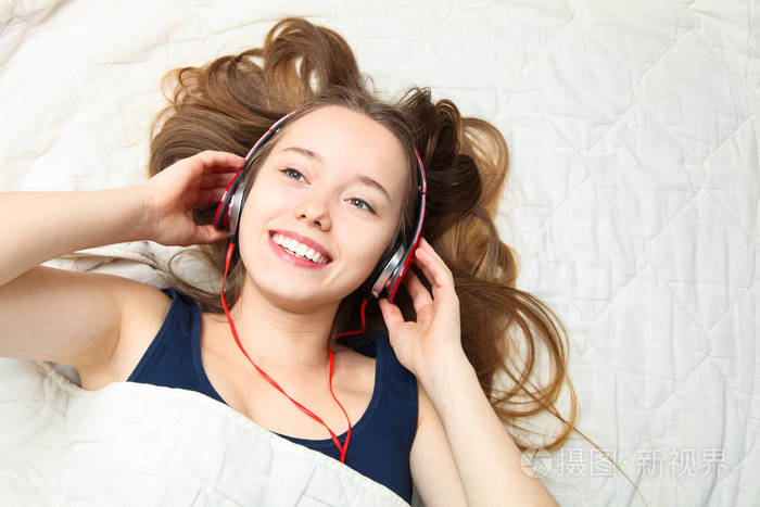 年轻美丽快乐的白种人女孩躺着在白色的床上用耳机听