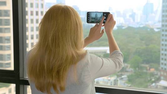 优雅的金发碧眼的女人在电话里拍照。女人从摩天大楼的窗户拍下了风景