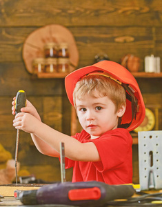 穿着橙色头盔和红色 t恤的男孩忙着工作。集中的孩子捆绑螺丝入木头。小木匠玩螺丝刀