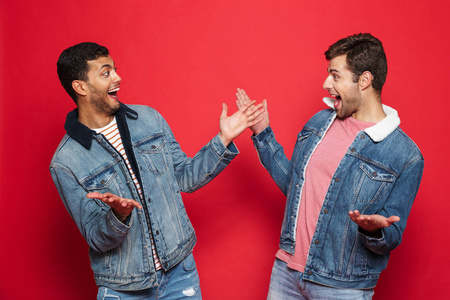 两个兴奋的年轻人朋友穿着牛仔夹克站在红色背景上，庆祝