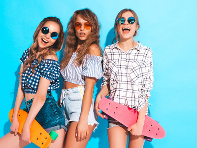 三个年轻时尚，，微笑，美丽的女孩与五颜六色的硬币滑板。 夏天的女人穿着时髦的格子衬衫，穿着太阳镜隔离在蓝色墙上。 积极的模特