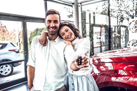 冒险正在等待。 美丽幸福的年轻夫妇拥抱着他们的新车钥匙，在经销商处愉快地微笑着
