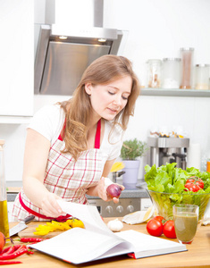 穿围裙的漂亮年轻女人在厨房里读食谱和做蔬菜沙拉