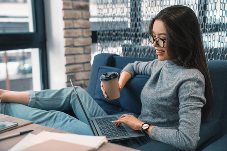 微笑的商务女性坐在沙发上，拿着纸杯咖啡在笔记本电脑上工作