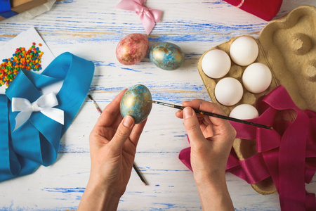 女性在画复活节彩蛋时的手，丝带，五颜六色的颜色，礼物放在白色的木桌上。
