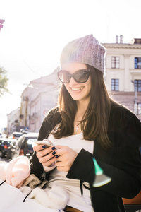 一个美丽的黑发女孩戴着太阳镜一顶针织帽和一个迷人的可爱微笑走在欧洲城市的街道上，购物时，一个白色的大纸板包裹，头发在她手一部智