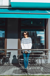美丽的黑发女孩戴太阳镜，舒适的针织套衫摆在城市咖啡馆的露台上