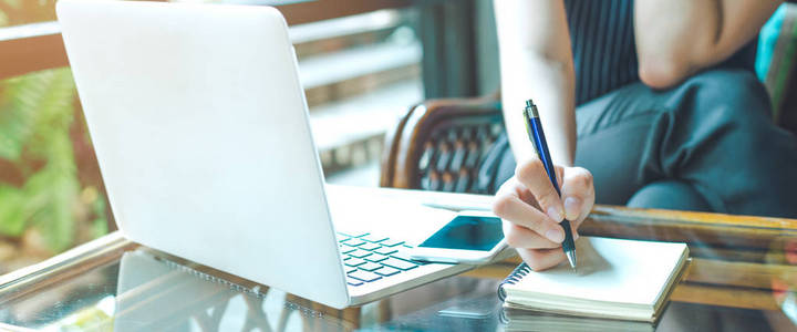商务女性的手是用钢笔写在记事本上，用笔记本电脑。网页横幅。