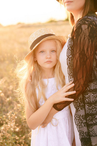 一个穿着白色连衣裙，戴着草帽，金发碧眼的女孩和一个姐姐在阳光明媚的一天日落时分在田野里