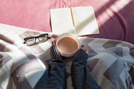 在懒洋洋的阳光明媚的早晨在床上喝咖啡和看书。女性双手捧着热气腾腾的热可可在床上看书