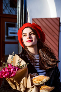 年轻漂亮的女法国女人，身穿红色贝雷帽和黑色外套，带着一束鲜花和法式小面包沿着欧洲城市的街道走。愉快的微笑和迷人的外表，法国式的巴