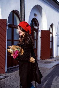 年轻美丽的女孩，法国女人，黑发，穿着红色贝雷帽和黑色外套，沿着欧洲城市的街道，带着一束一束鲜花和法国小面包。 愉快的微笑和迷人的