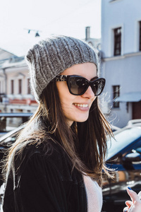 一个美丽的黑发女孩戴着太阳镜一顶针织帽和一个迷人的可爱微笑走在欧洲城市的街道上，购物时，一个白色的大纸板包裹，头发在她手一部智
