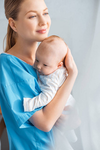 美丽快乐的年轻母亲抱着可爱的婴儿，望着医院的房间