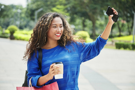 漂亮的印尼女性，带着一杯热饮料，微笑着，用智能手机挥动着手，在购物时站在模糊的街道背景上