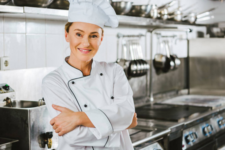 有魅力的女厨师穿着制服，双臂交叉，看着餐厅厨房里的摄像机