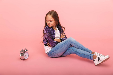 一个心烦意乱的小女孩孤零零地坐在粉红色的背景下，正拿着闹钟