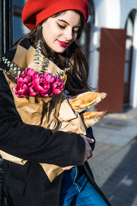 年轻美丽的女孩，法国女人，黑发，穿着红色贝雷帽和黑色外套，沿着欧洲城市的街道，带着一束一束鲜花和法国小面包。 愉快的微笑和迷人的