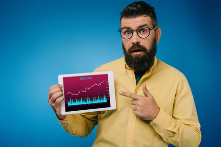 震惊的胡须男子指向数字平板电脑与塔利丁应用程序，孤立在蓝色。