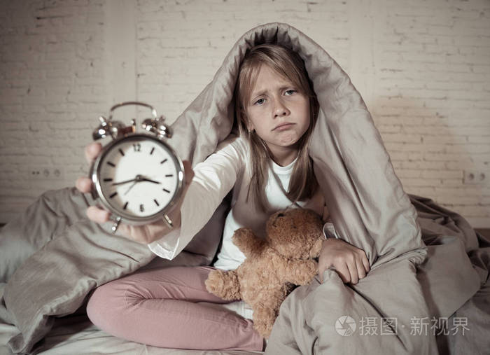 可爱的失眠小女孩坐在床上,闹钟看起来很累,有睡眠问题,晚上睡觉,或者
