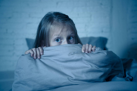 害怕的小女孩睡不着,躲在羽绒被后面,在黑暗中惊恐地看着,做着童年