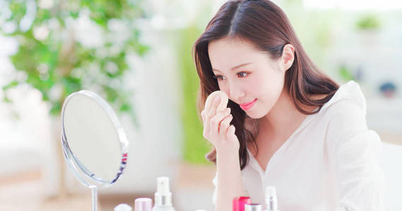 美丽的亚洲女人在脸上用化妆海绵，在家里照镜子