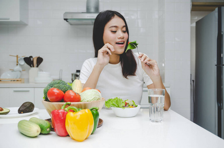 美丽的年轻亚洲女人苗条的身体穿着白衬衫节食和吃健康的食物新鲜蔬菜沙拉坐在厨房内部的家庭生活方式，良好的健康饮食，食物和饮料的概念