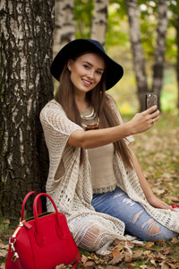 在秋天的森林里，一位戴着帽子的年轻美女坐在手机上拍照